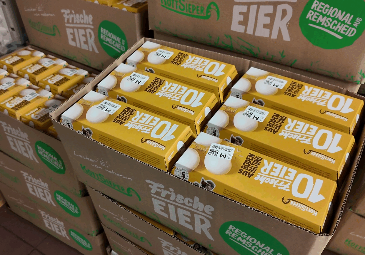 Stapel von gelben Kartons mit der Aufschrift "10 frische Eier" sind in Pappkartons verpackt, die für die Lebensmittelbranche fein säuberlich angeordnet sind. Einige Kartons sind mit grünen Etiketten versehen, die auf regionale Produkte hinweisen.