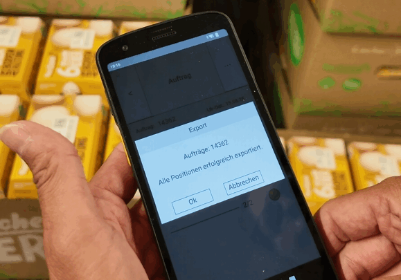 Eine Person hält ein Smartphone in der Hand, auf dem eine Exportbestätigung in deutscher Sprache angezeigt wird, während im Hintergrund Regale mit verpackten Eiern zu sehen sind - eine Anspielung auf die Digitalisierung der Lebensmittelbranche und ERP für Lebensmittelproduzenten. Das Telefon zeigt Optionen zum Bestätigen oder Abbrechen an.